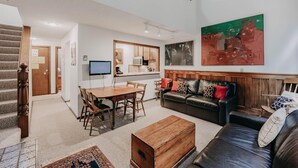 A spacious living area with a wooden dining table, a leather sofa, and a coffee table. A kitchen is visible in the background with a microwave and a TV. - Snowater Condo #57 - A Great 2-story Family Condo That Sleeps 6! (Deming)