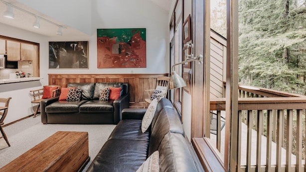 A modern living room with a black leather sofa, wooden coffee table, and a dining area with a wooden table and chairs. There is a large painting on the wall and a view of a forest through a balcony. - Snowater Condo #57 - A Great 2-story Family Condo That Sleeps 6! (Deming)