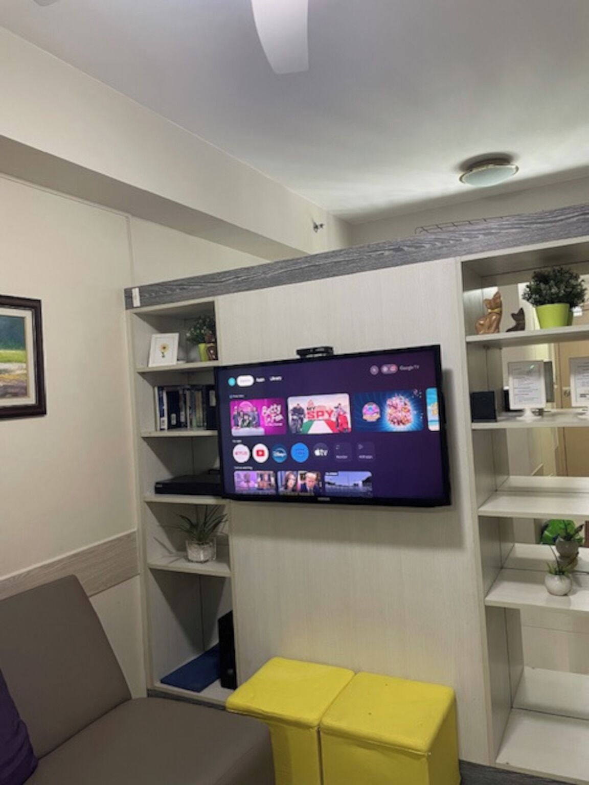 A living room with a flat-screen TV mounted on the wall, a sofa, a yellow ottoman, and shelves with various items.