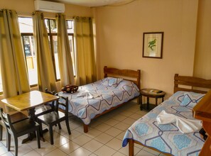 Two single beds with matching headboards, a wooden table and chair, and a small table with a chair, all set against a backdrop of a window with curtains.