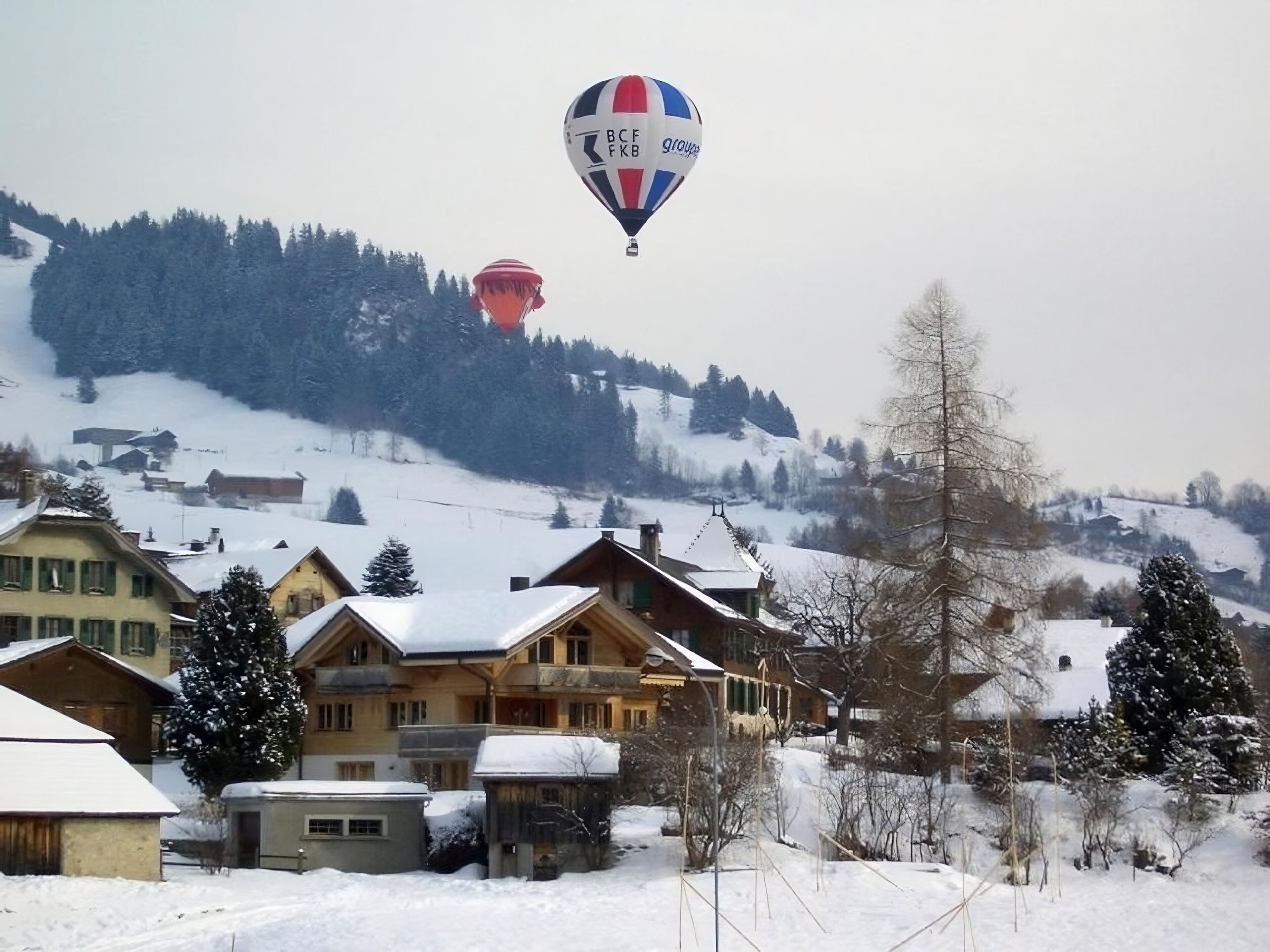 Un village enneigé, avec des maisons traditionnelles en bois, et un ballon à air chaud en arrière-plan.