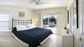 Quarto com cama, ventilador de teto, janela com persianas, porta e vista para área de piscina externa.