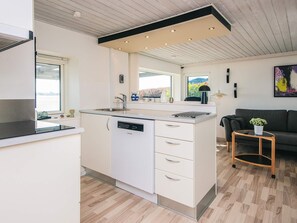 A modern kitchen with a dishwasher, a sink, and a window. There is a small table with a plant and a couch in the living area.