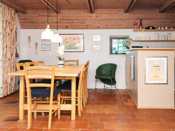 A dining area with a wooden table and chairs, a green chair, a small island with a sink, and a window with a view of greenery.