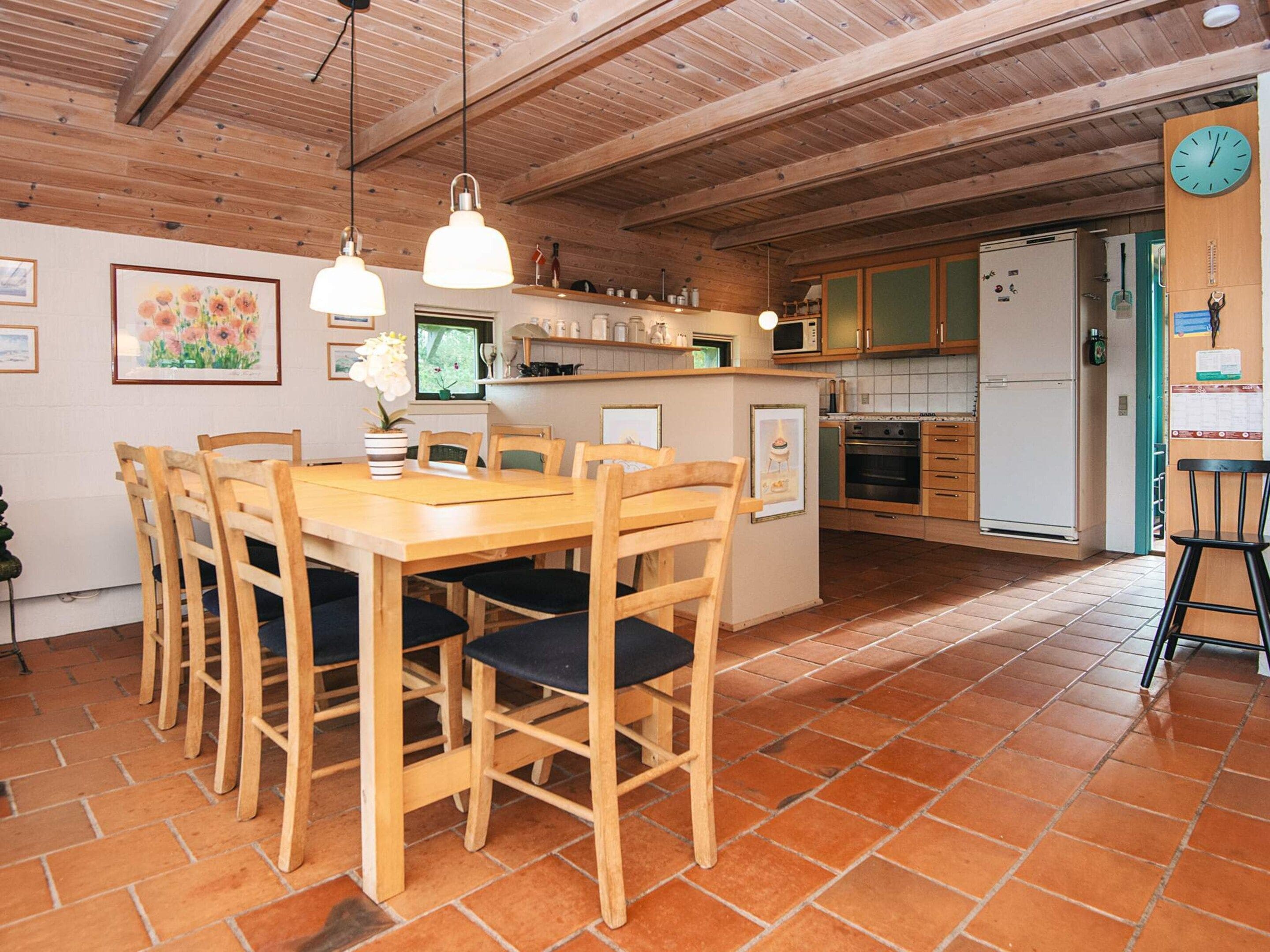 A wooden dining table with chairs, a kitchen area with cabinets and appliances, and a clock on the wall.