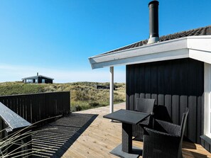 A wooden deck with a table and chairs, a black chimney, and a modern house in the background.