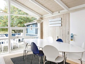 Comedor con mesa y sillas blancas, techo de madera y vista a una casa azul a través de amplios ventanales.