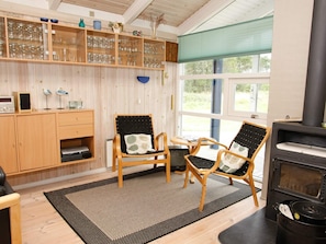 Une pièce chaleureuse avec un plancher en bois, deux fauteuils en osier dotés de coussins, une bibliothèque contenant de la verrerie, une radio et un poêle à bois.