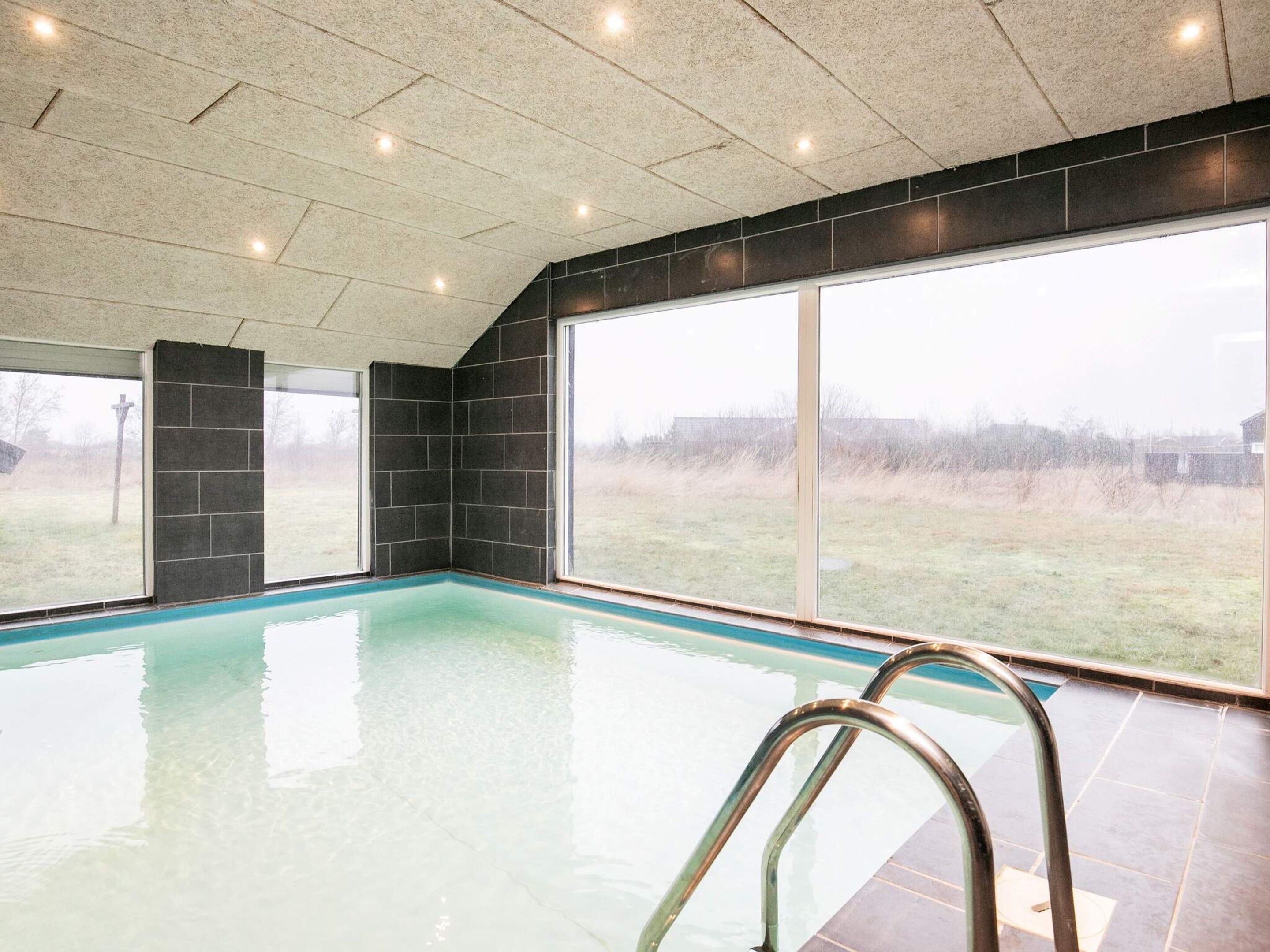 An indoor swimming pool with clear water, surrounded by large windows offering a view of the outdoors.
