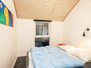 A bedroom with a sloped ceiling, a window, a bed with blue bedding, a white pillow, and a wall-mounted lamp.