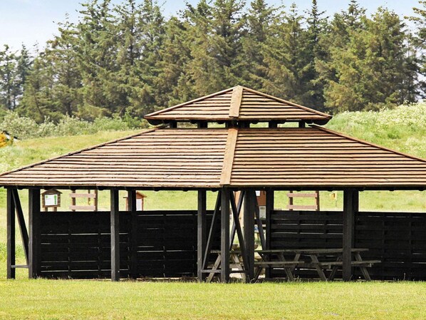 A wooden pavilion with a sloped roof, supported by black beams, set in a grassy field with trees in the background.