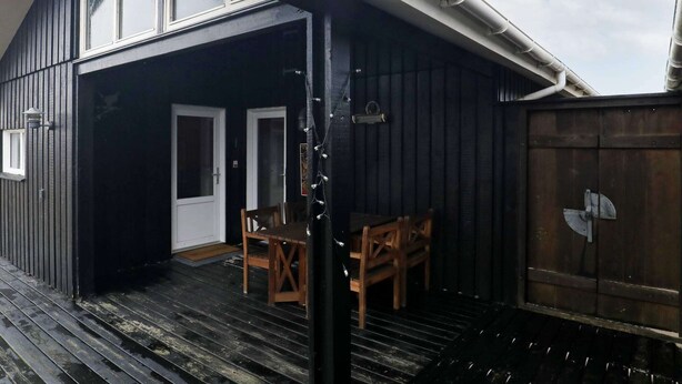 Een overdekt buitengebied met een houten terras, twee deuren en een kleine tafel met stoelen.