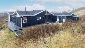 Een rij zwarte blokhutten met schuine daken en een houten terras, gelegen in een grasrijke omgeving onder een heldere lucht.