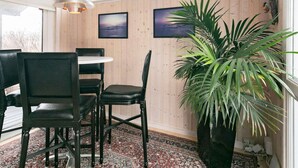Een kamer met een houten lambrisering, een ronde eettafel met stoelen, een grote potplant en twee ingelijste foto's.