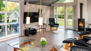 A modern living room with a fireplace, comfortable seating, a flat-screen TV, and large windows offering a view of the outdoors. - Luxury Spa Retreat - By Traum Ferienwohnungen (Glesborg)
