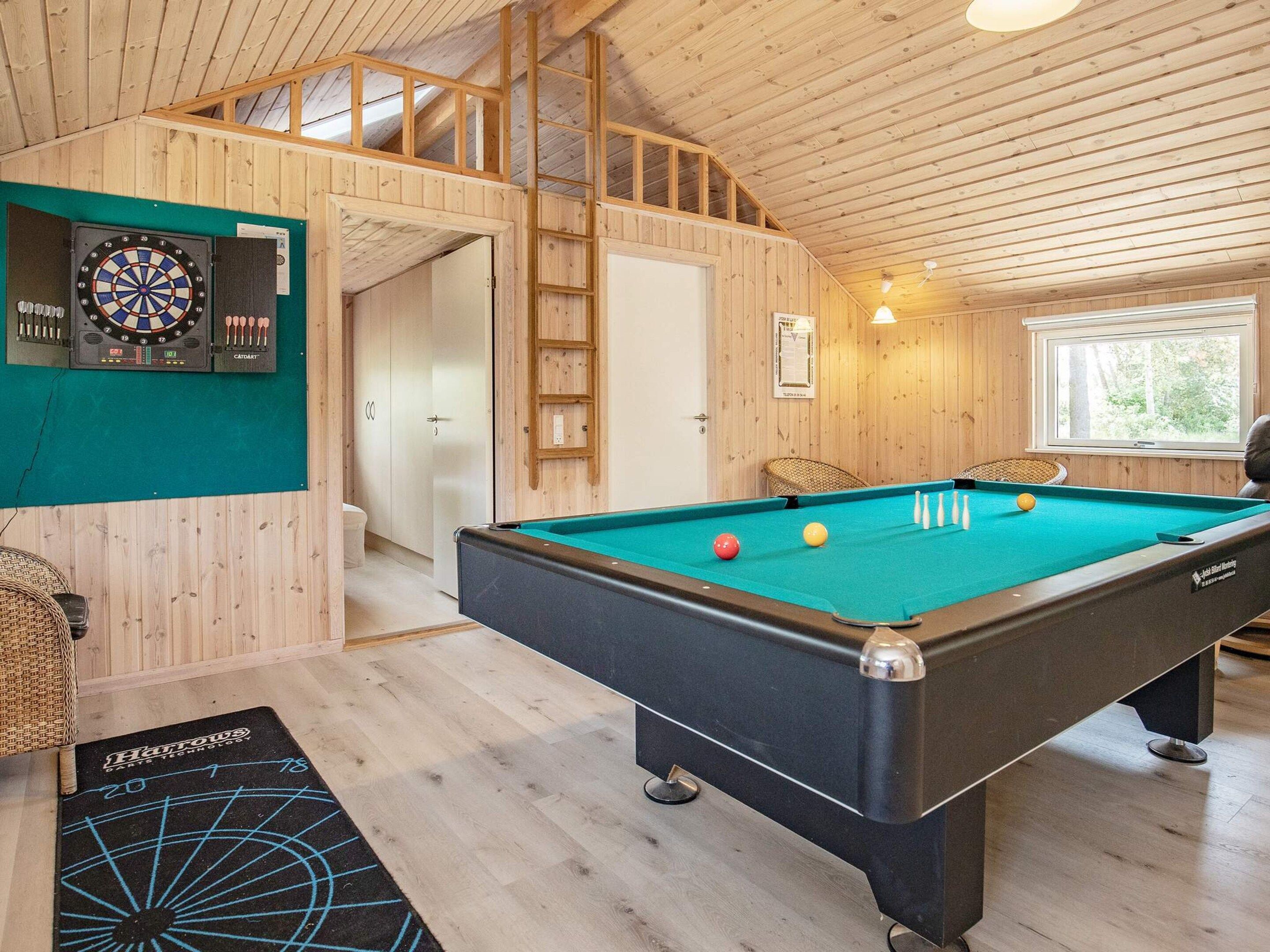 Ein Billardtisch mit Billardkugeln, eine Dartscheibe und ein Billardköchengestell in einem Raum mit Holzwänden und einer geneigten Decke.