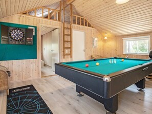 A pool table with billiard balls, a dartboard, and a billiard cue rack in a room with wooden walls and a sloped ceiling.
