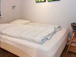 A single bed with a white mattress and a striped blanket, a wooden chair, and a small table with a patterned cloth.