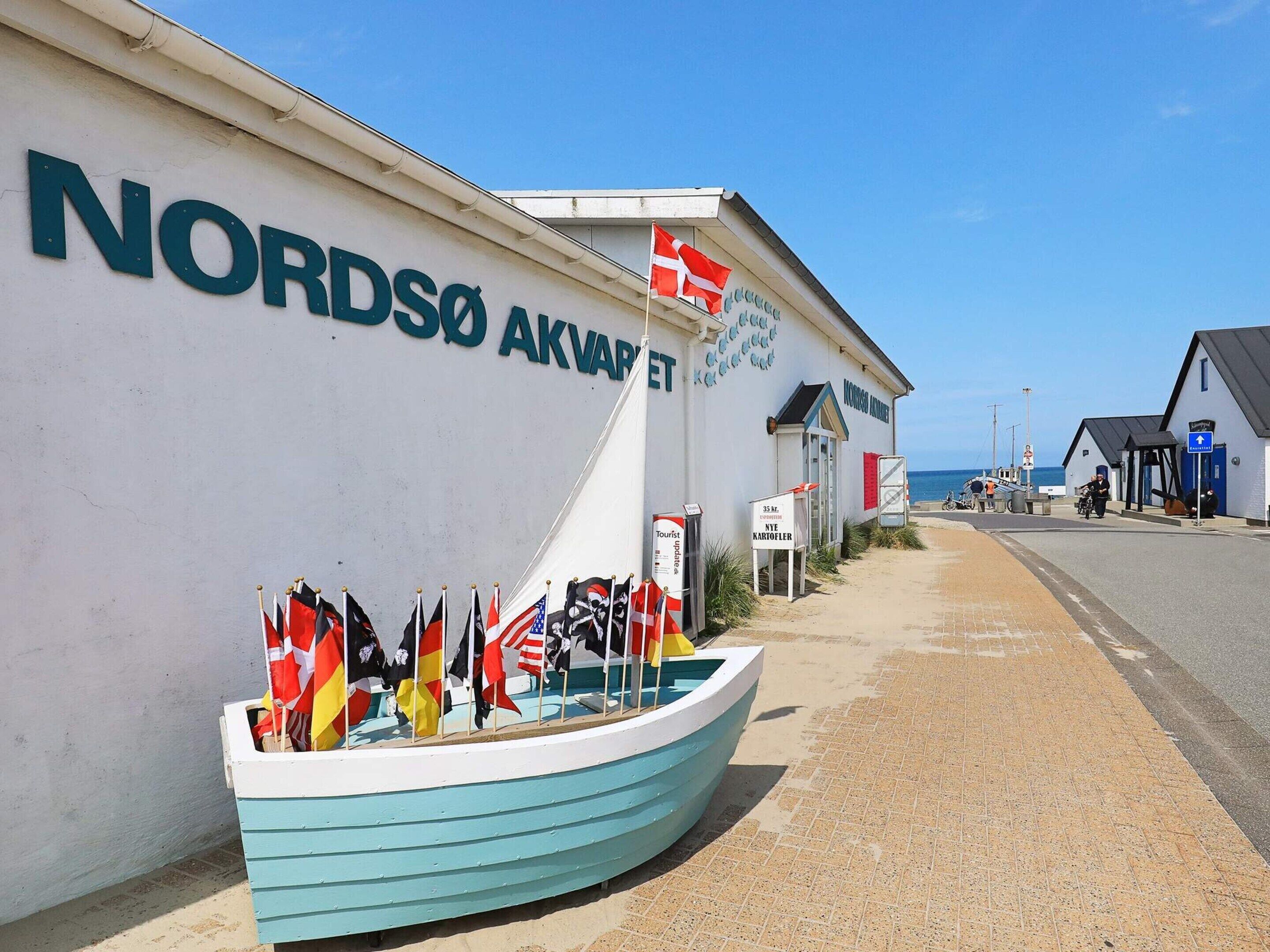 A building with a sign reading "NORDSØ AKVAPET" and a boat with various flags in front.