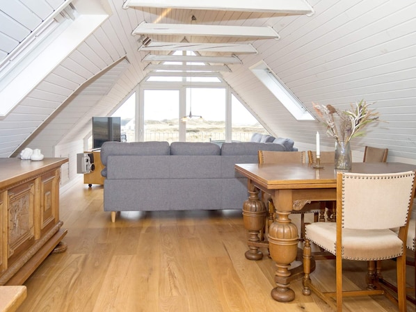A loft-style room with a wooden floor, a grey sofa, a wooden dining table with chairs, and a TV on a wooden cabinet.