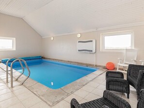 En indendørs swimmingpool med en stige, omgivet af fletningsmøbler og et vindue.