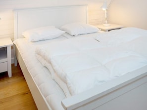 A neatly made bed with white bedding and pillows, a bedside table with a lamp, and a wooden floor.