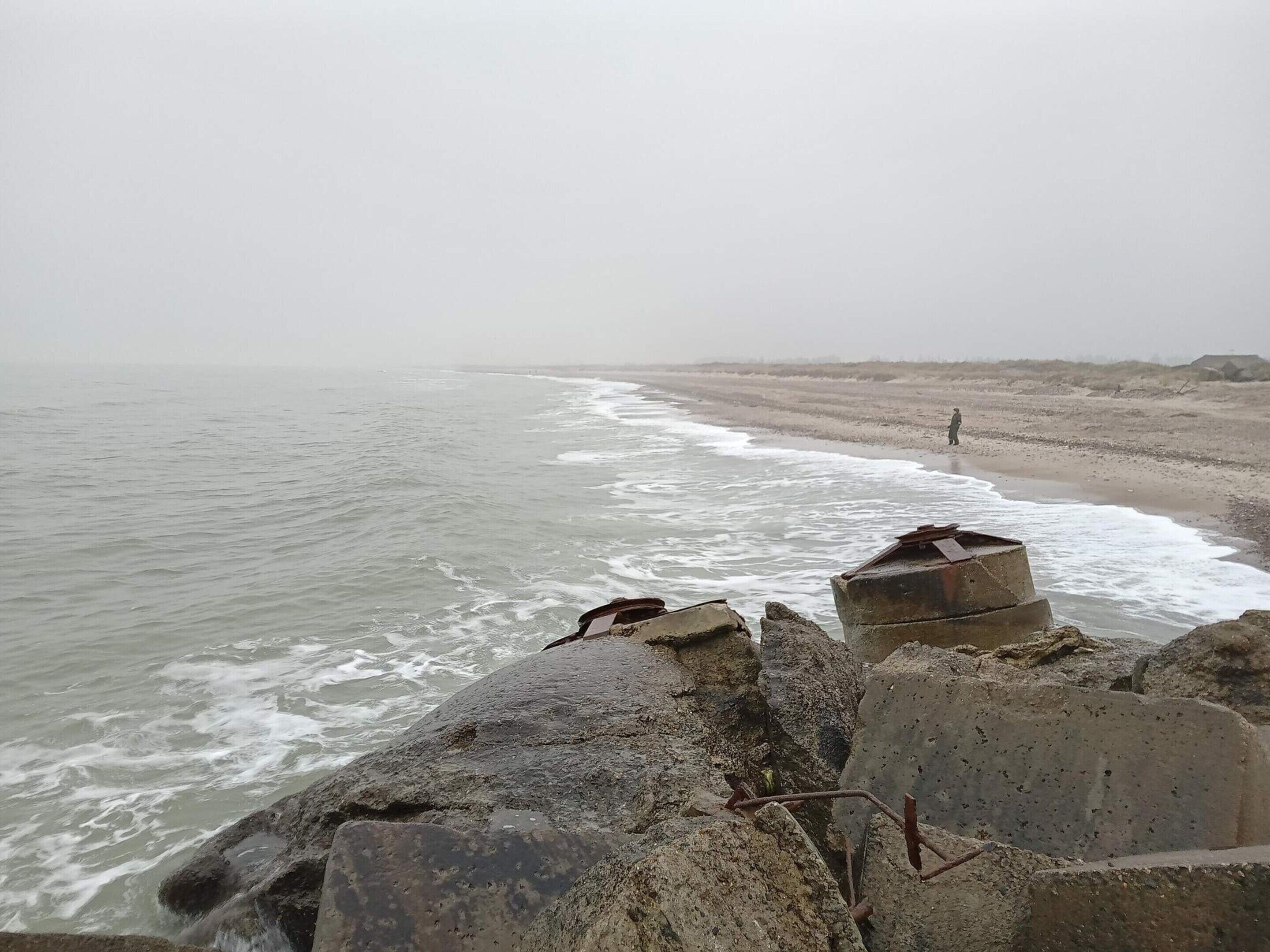 A coastal scene with a rocky foreground, a sandy beach, and a person standing in the distance.