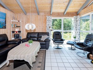 A modern living room with a black leather sofa, a dining table with a patterned tablecloth, and a large window offering a view of greenery. - 4 Star Holiday Home in Fjerritslev (Fjerritslev)