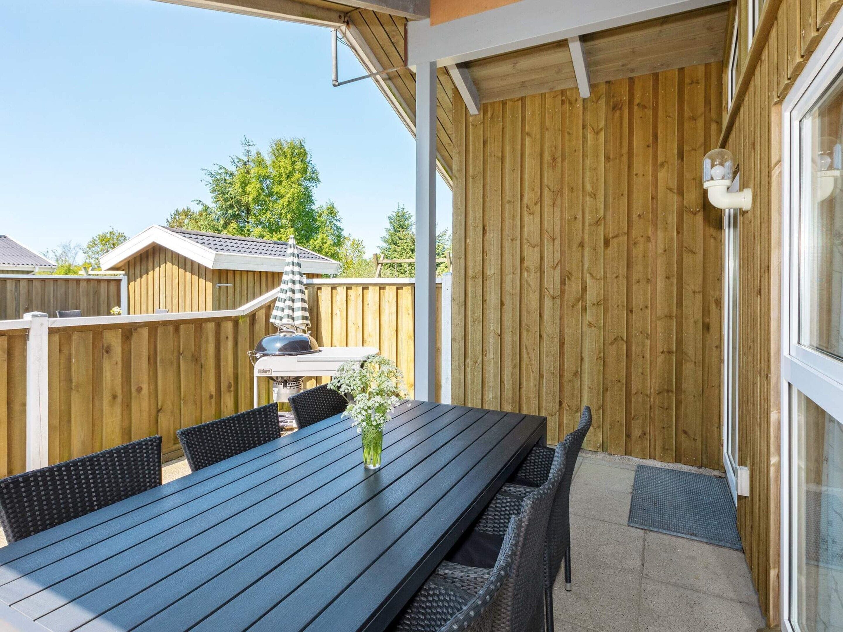 Une terrasse extérieure avec une longue table et des chaises, un barbecue et une clôture en bois.