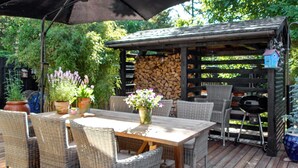 An outdoor patio with a wooden table and wicker chairs, surrounded by potted plants and a barbecue grill. - Attractive Holiday Home in Hemmet near Sea (Hemmet)