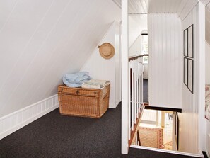 Une pièce avec un plafond en pente, un panier en osier rempli de vêtements, un chapeau accroché au mur et une vue sur un couloir orné d’un tapis à motifs.