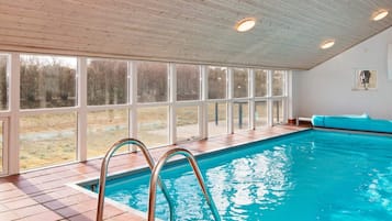 Una piscina interior con vistas despejadas al exterior gracias a amplios ventanales, suelo de baldosas y techo de madera.