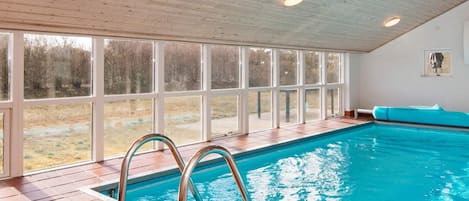 An indoor swimming pool with a clear view of the outdoors through large windows, a tiled floor, and a wooden ceiling.