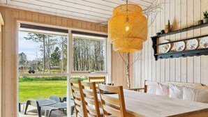 Une table à manger en bois, des chaises, un luminaire suspendu et une vue sur un espace verdoyant grâce à de grandes fenêtres.