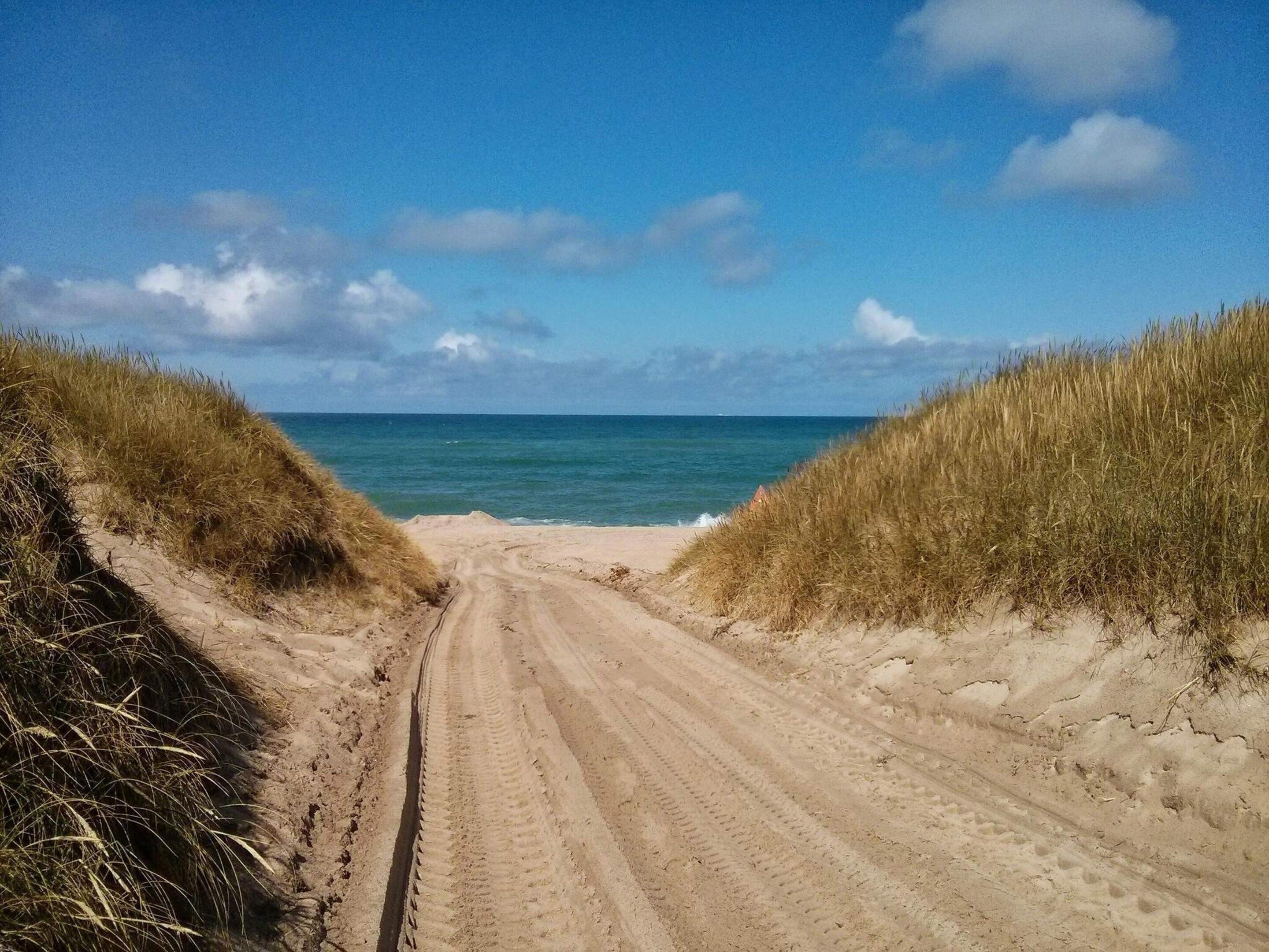 Ein sandiger Pfad führt zum Strand, gesäumt von grasbewachsenen Dünen.