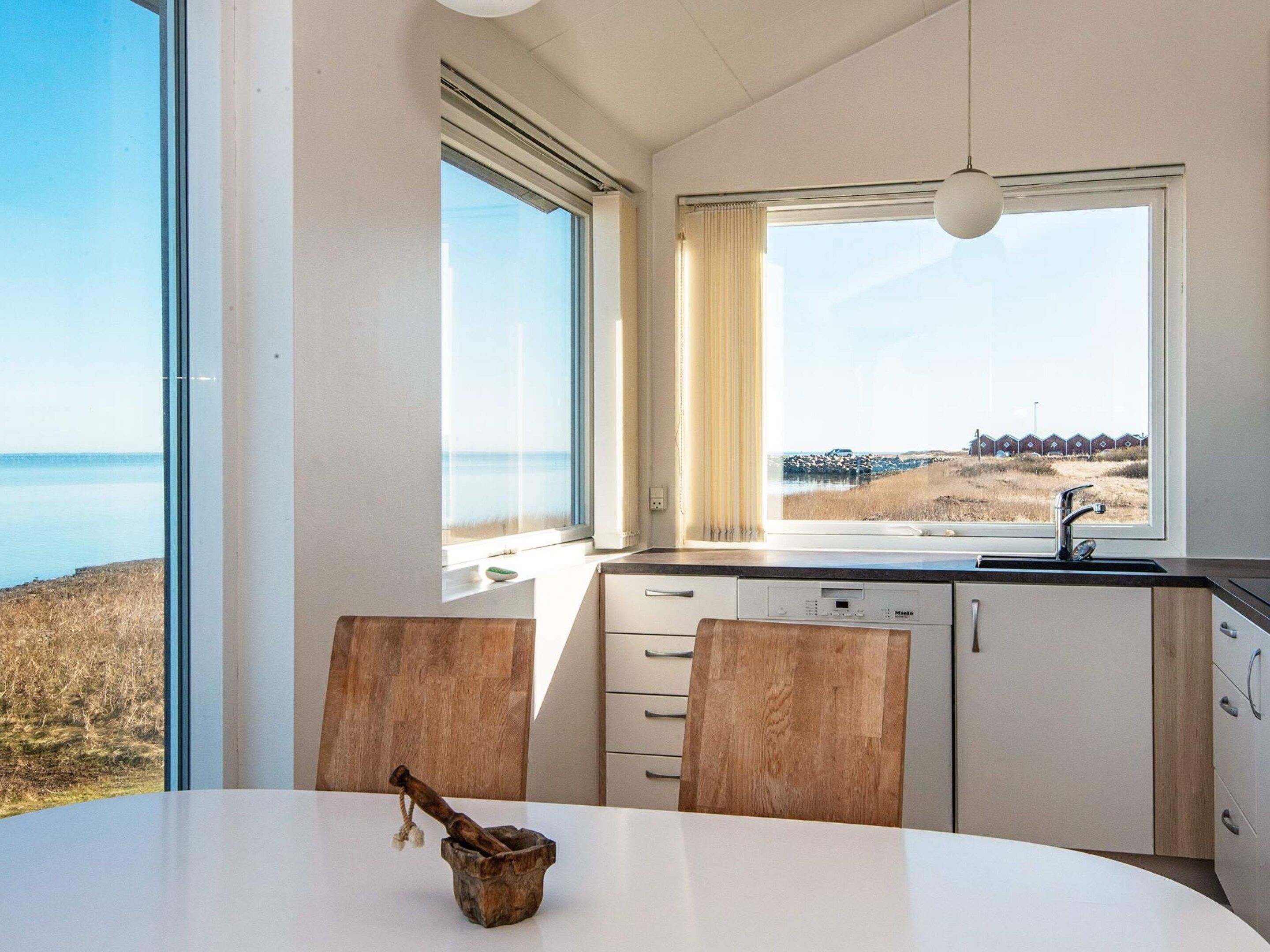 A modern kitchen with a view of the sea, featuring a dining table, wooden chairs, and a built-in dishwasher.