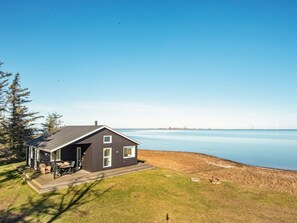 A house with a dark exterior, a covered patio with outdoor furniture, and a view of a calm body of water and distant wind turbines. - 4 Star Holiday Home in Harboore (Harboore)