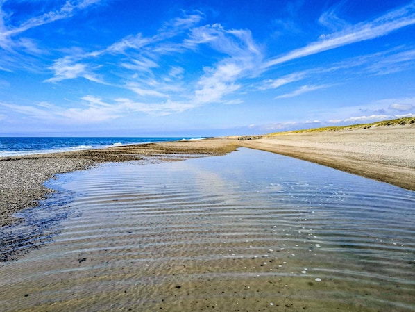 En strand med klart vatten, en sandstrand och en avlägsen vy över havet under en blå himmel.