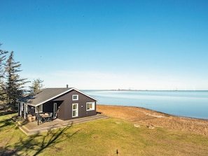 A house with a dark exterior, a covered patio with outdoor furniture, and a view of a calm body of water and distant wind turbines.
