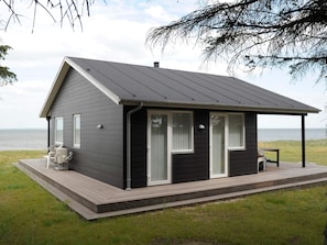A small, dark-colored cabin with a deck, two windows with white frames, and a view of the water.