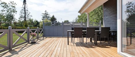 Una terraza de madera con mobiliario exterior, entrada protegida por un portón y vistas a árboles y al cielo.