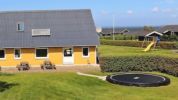 A residential building with a grey roof, a yellow facade, and a satellite dish. There is a playground with a slide and a swing set. A trampoline is visible in the yard.