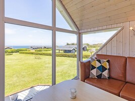 Una habitación con un ventanal grande, techo de madera, un sofá de cuero marrón con un cojín estampado y una pequeña mesa con una vela.