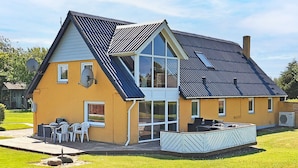 Une maison à deux étages, dotée d’un toit en pente, de grandes fenêtres et d’un patio aménagé avec du mobilier d’extérieur.