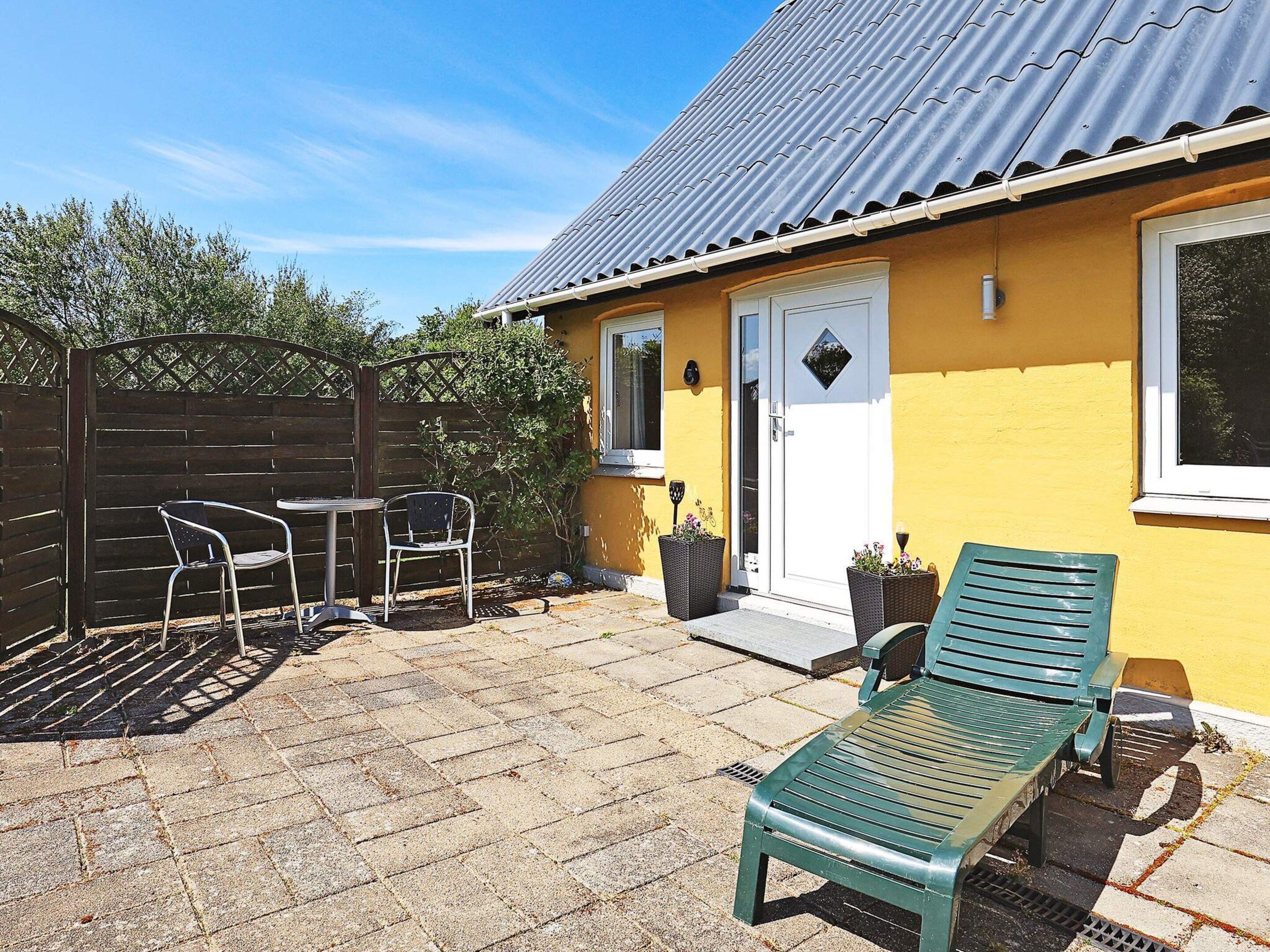 A yellow building with a tiled roof, a door, and a window. There is a paved patio with a green bench and two chairs.