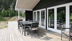 A deck with a table and chairs, a covered area with a sloped roof, and a glass door leading to a garden. - 4 Star Holiday Home in Albaek (Aalbaek)
