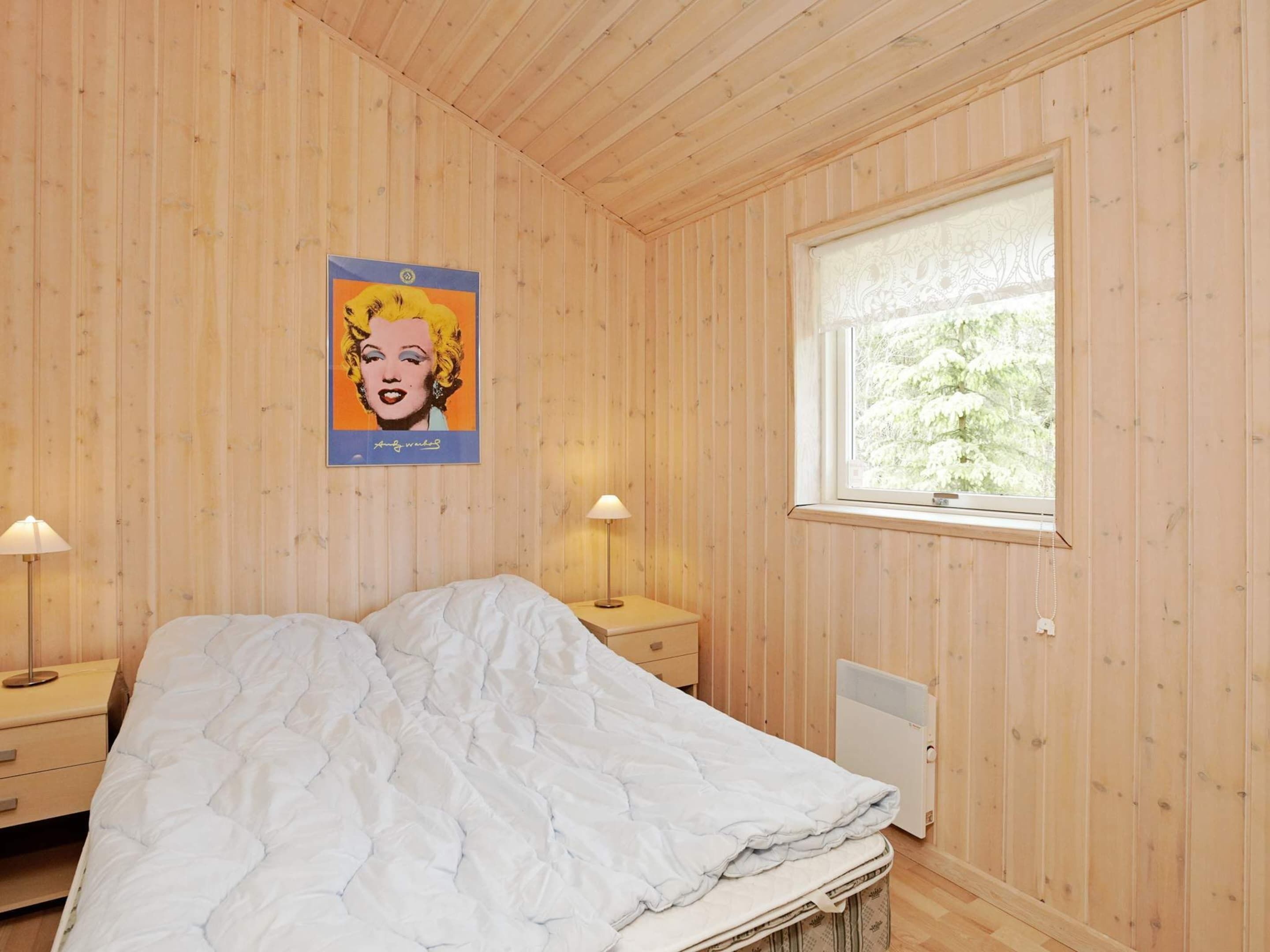 Ein Schlafzimmer mit Holzwänden, einem Bett mit weißer Tagesdecke, zwei Nachttischlampen, einem Heizkörper und einem Fenster mit Blick auf Bäume.