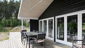 En terrasse med et bord og stole, et overdækket område med skråt tag og en glasdør, der fører til en have.