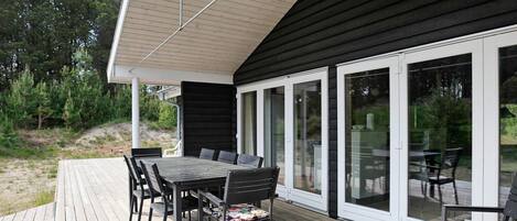 A deck with a table and chairs, a covered area with a sloped roof, and a glass door leading to a garden.