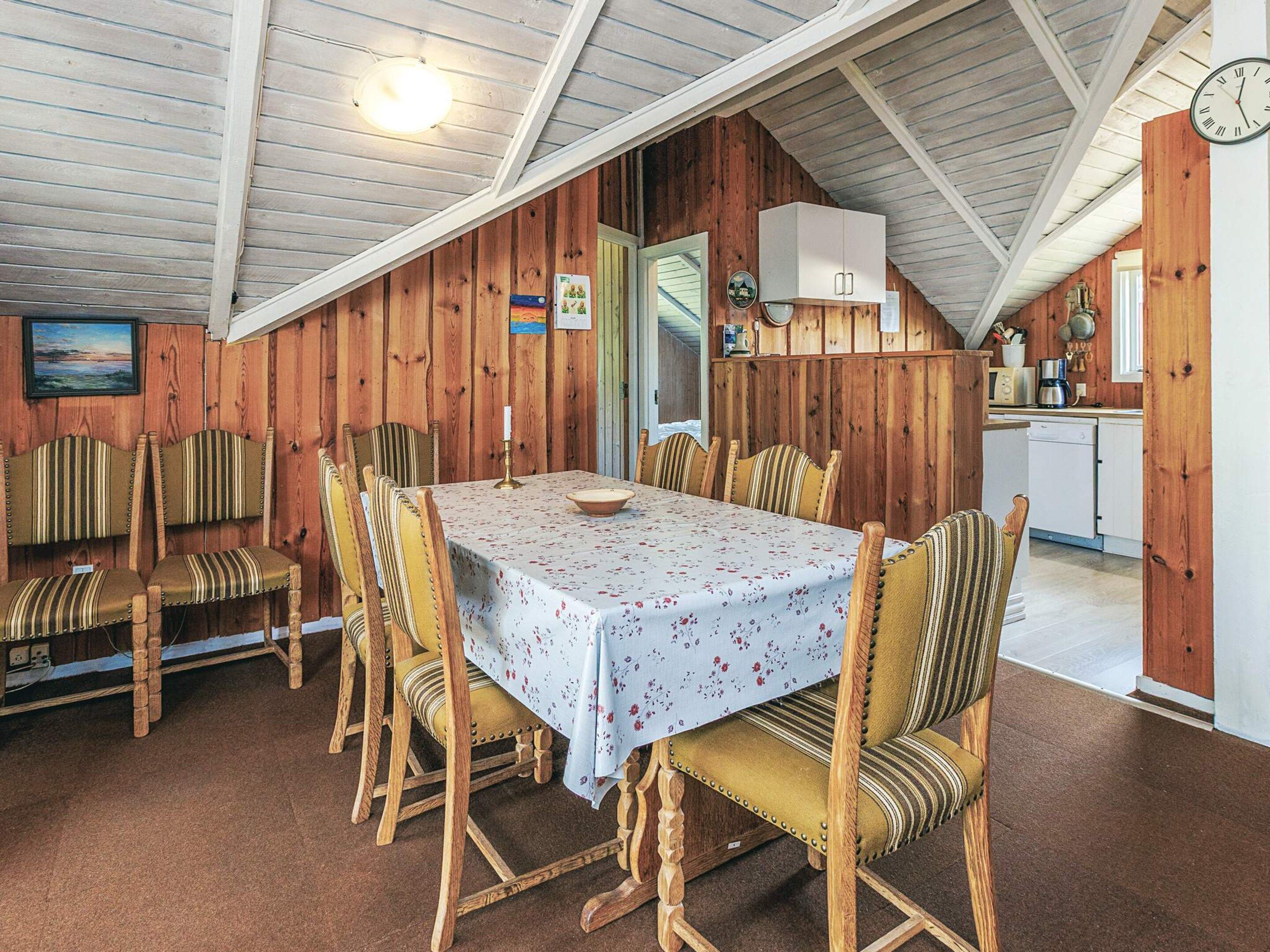 A dining area with a table set for six, wooden chairs, a clock, and a kitchen in the background.